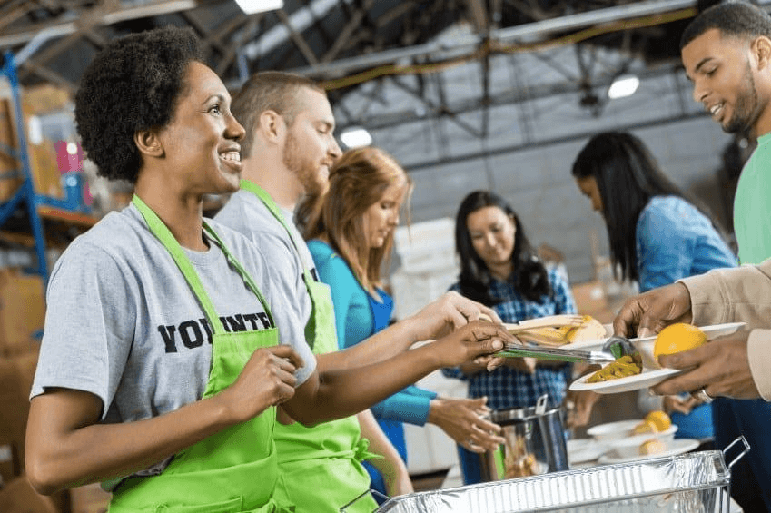 mulher negra serve refeição para uma pessoa, ela usa avental verde e camiseta cinza