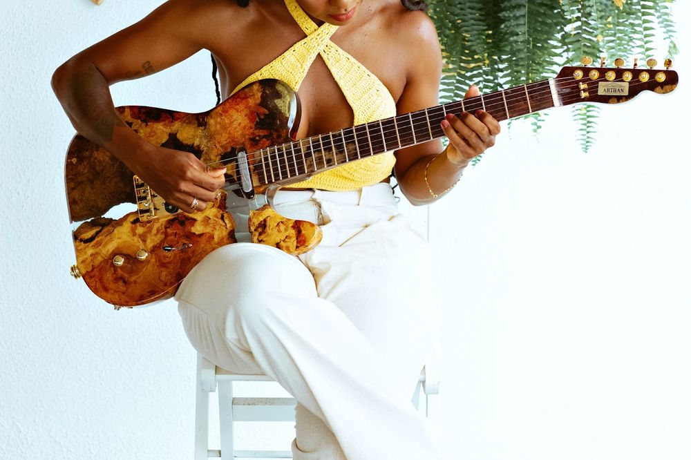 Mulher negra de blusa amarela, cabelo trançado e calça branca tocando guitarra 