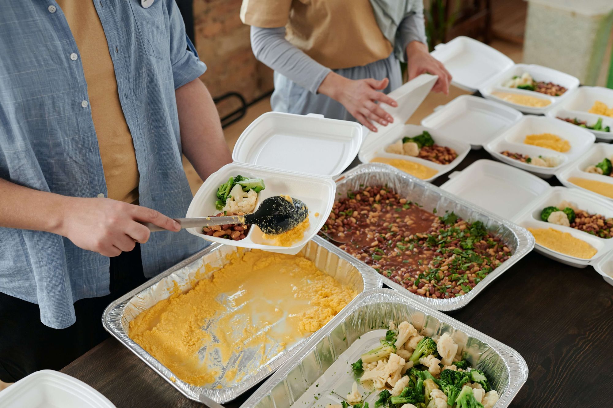 A imagem mostra duas pessoas próximas a uma mesa onde há diversos recipientes descartáveis com alimentos preparados. Elas montam marmitas de isopor com os alimentos.