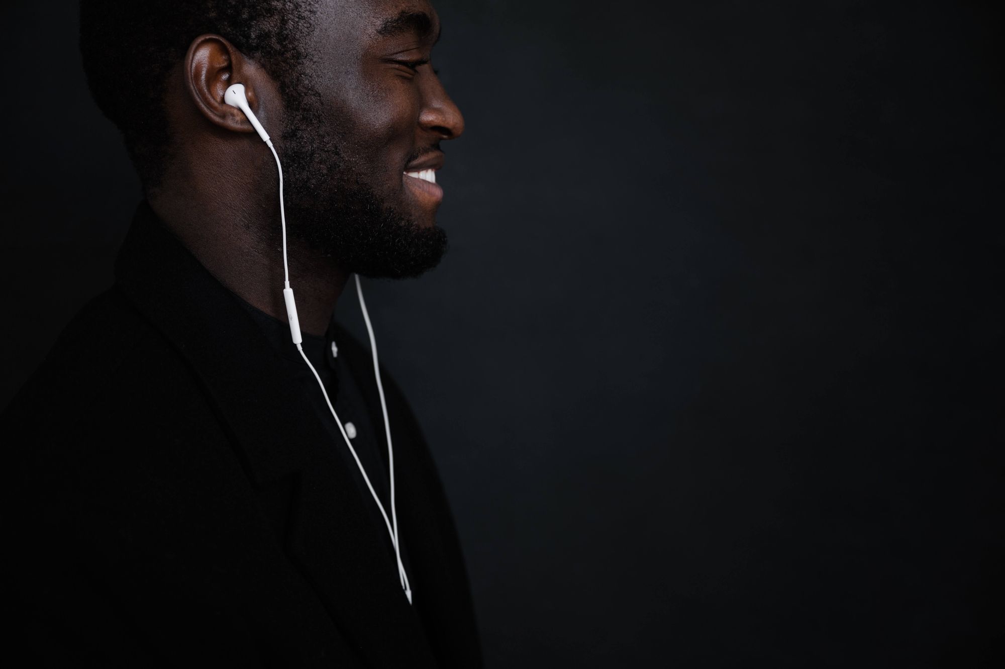 homem negro em parede preta, de perfil, sorrindo com fones de ouvido branco no ouvido 