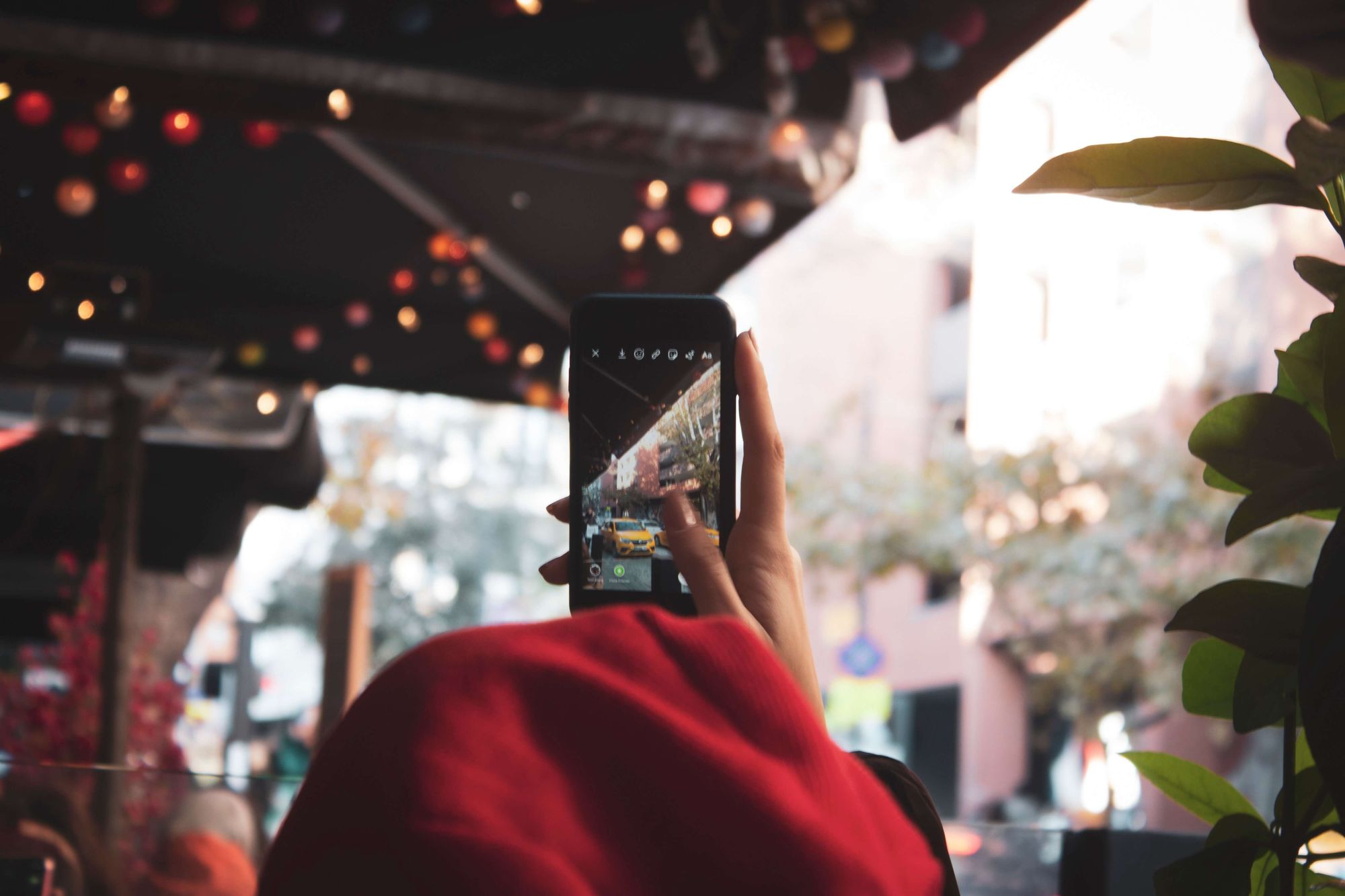 Foto em detalhe de uma mão segurando um celular. Na tela do aparelho, uma captura de imagem da rua, por onde passam carros amarelos.