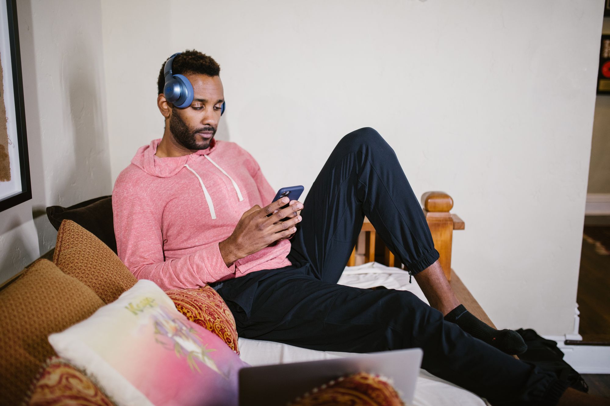 Foto de um homem negro de cabelos curtos segurando um celular com as duas mãos. Ele está sentado em um sofá e usa um fone de ouvidos um pouco acima de uma das orelhas. Usa casaco de moletom rosa e calça preta.