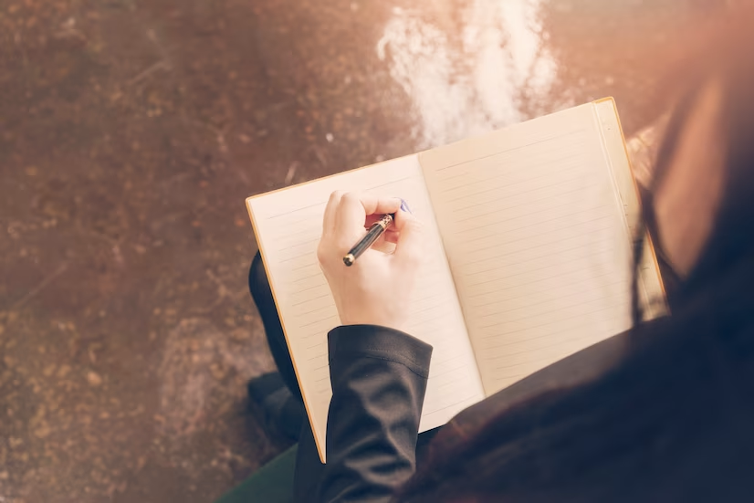 mão branca e feminina apoia caneta sob uma folha em branco de caderno em algum ambiente externo