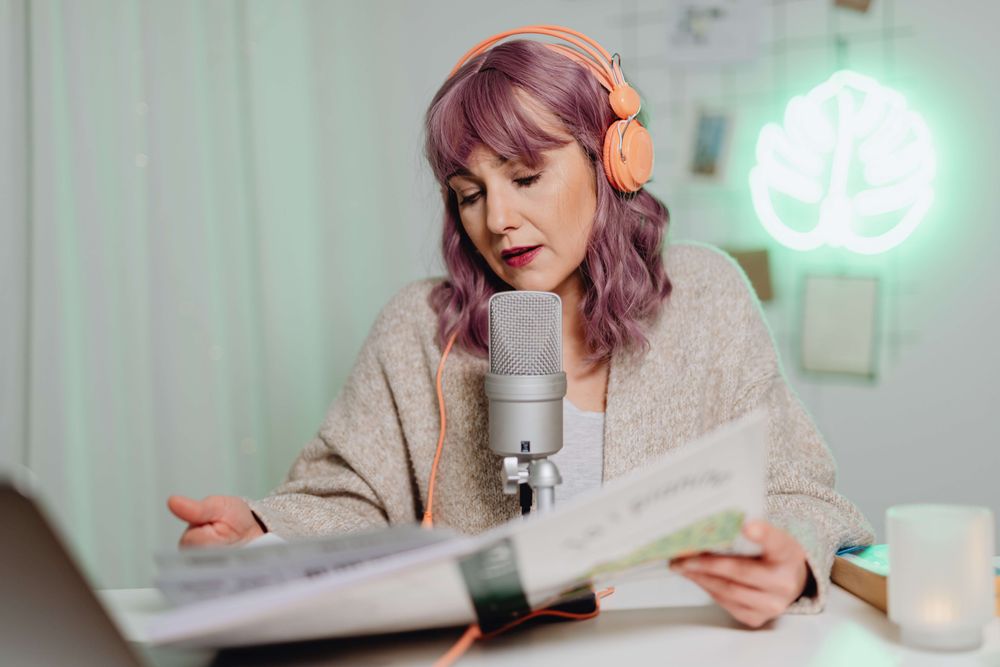 mulher de cabelo rosa com um fone de ouvido laranja e falando em um microfone profissional enquanto lê algo