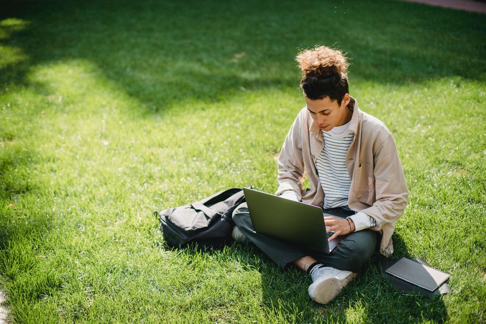 homem branco, de cabelo preso, sentado na grama assistindo algo na tela de um computador