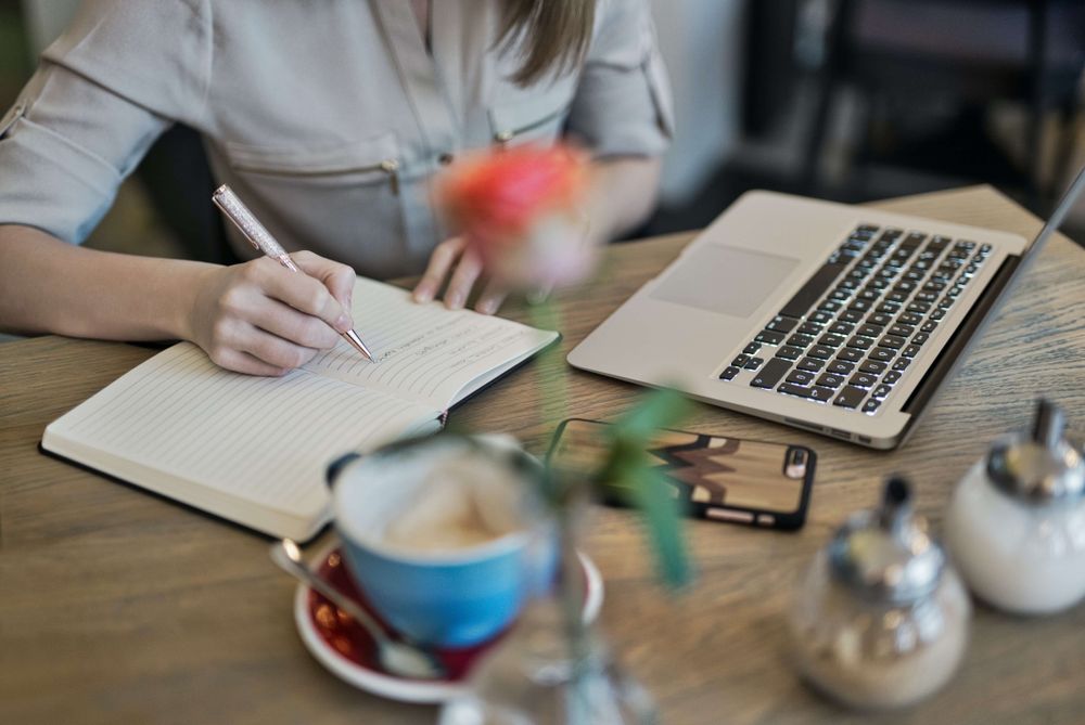 Mesa de trabalho com uma pessoa fazendo anotações em um caderno. Entre os objetos espalhados há uma xícara e um computador.
