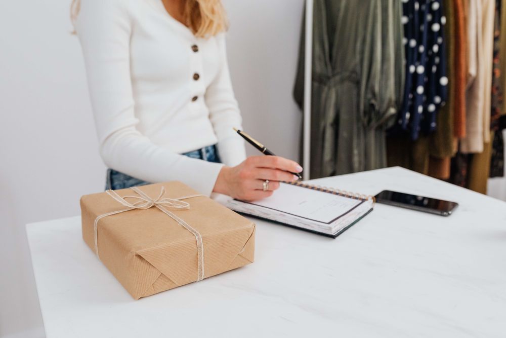 mesa com pacote embalado ao lado de um caderno em que uma mulher de camisa branca faz anotações