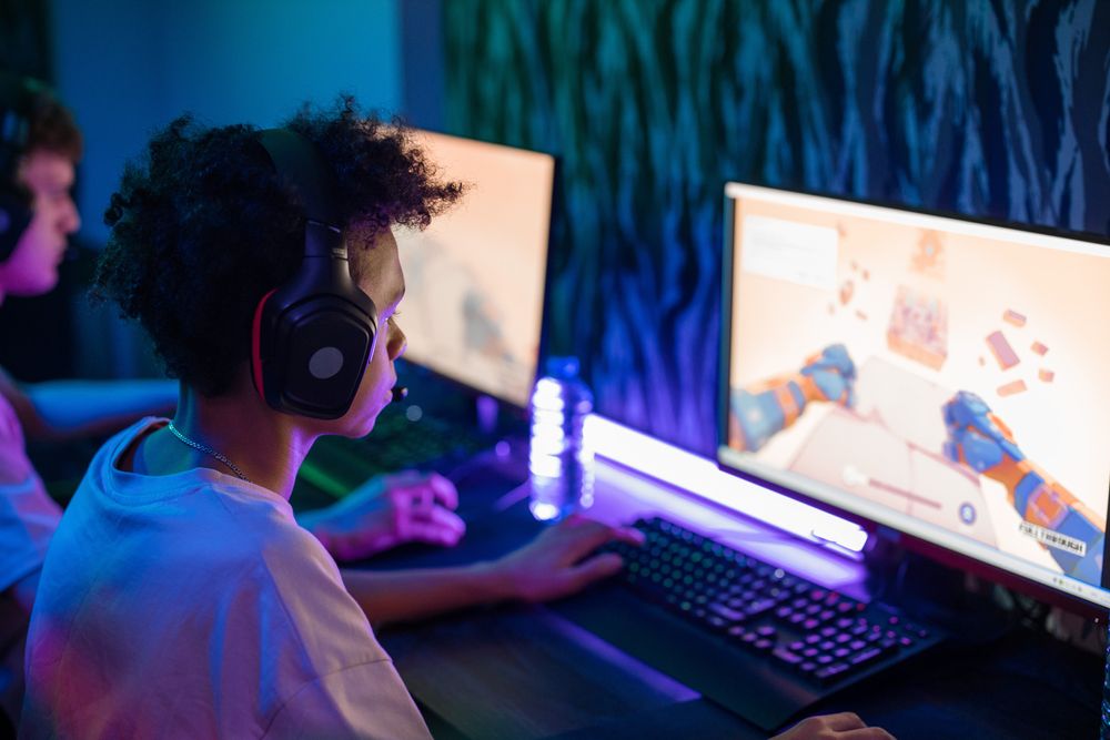 menino jogando no computador, com um fone de ouvido e ao lado de outro jovem gamer