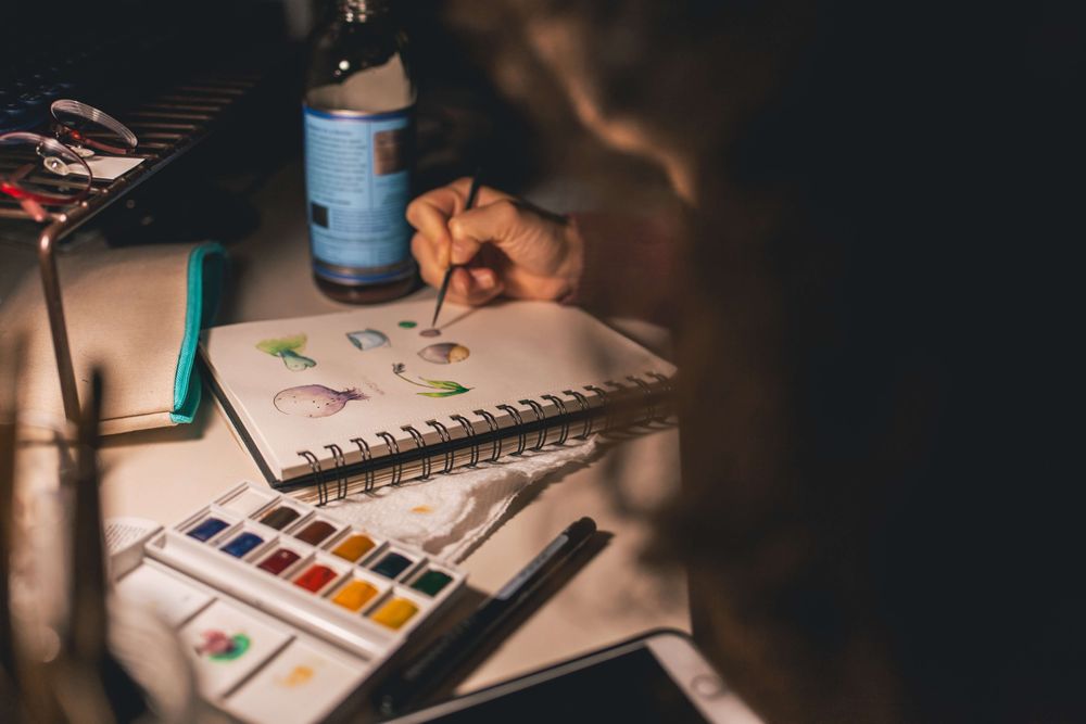 Mão desenhando em caderno sobre mesa com caixas de tintas.