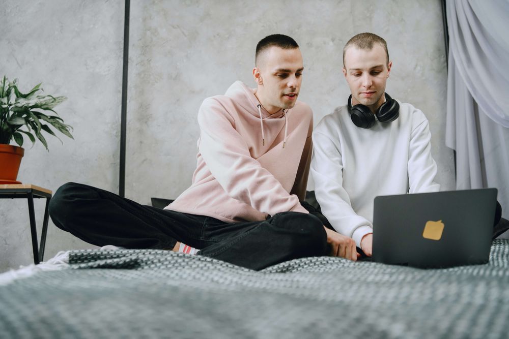 dois homens brancos e de cabelos raspados sentados em uma cama olhando para um notebook 