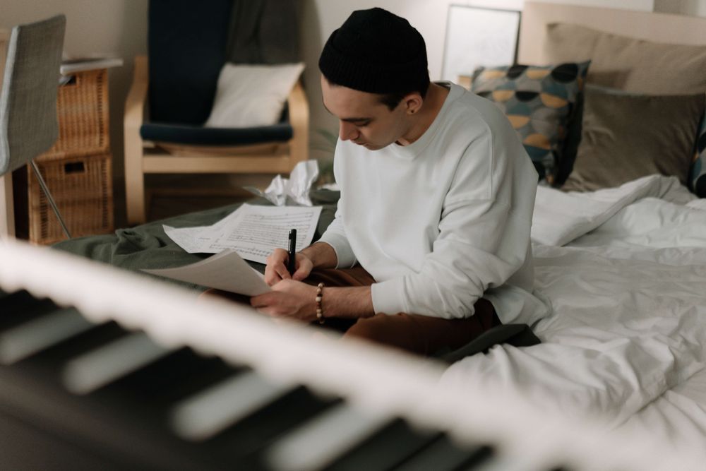 Homem sentado na cama escrevendo em folhas de papel. Em primeiro plano, um piano desfocado.