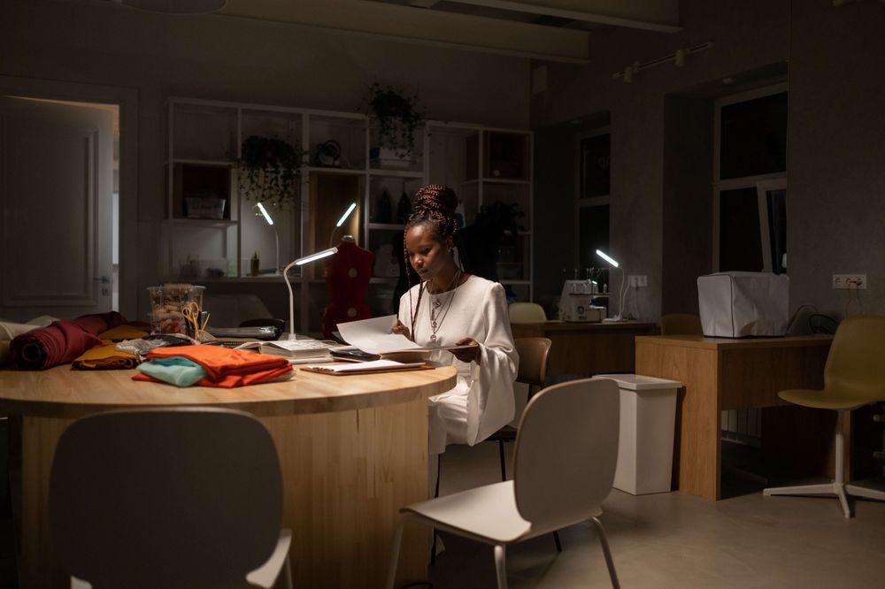 Foto de uma sala ampla de escritório com várias mesas. Ao centro há uma mulher negra mexendo em papeis.