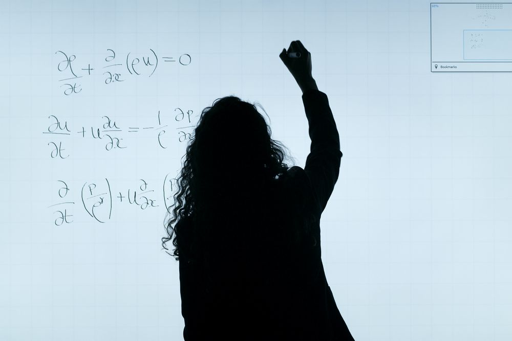 mulher contra a luz escrevendo em uma lousa branca onde há fórmulas matemáticas, vê-se somente a silhueta dela