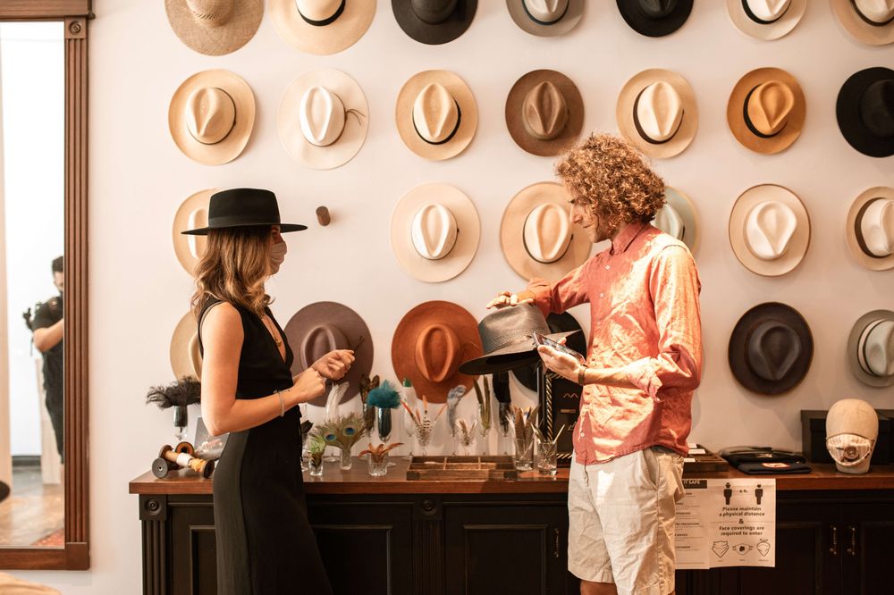 Homem e mulher de frente um para o outro, em uma loja de chapéus. Ele segura um chapéu na mão e ela observa. 