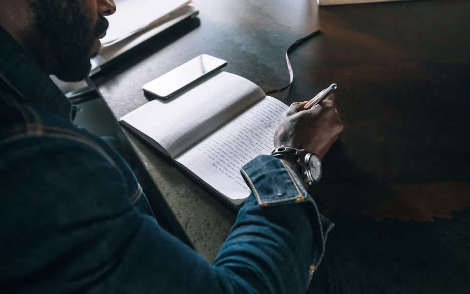 Homem negro vestindo jaqueta jeans e escrevendo em um caderno com celular ao lado.